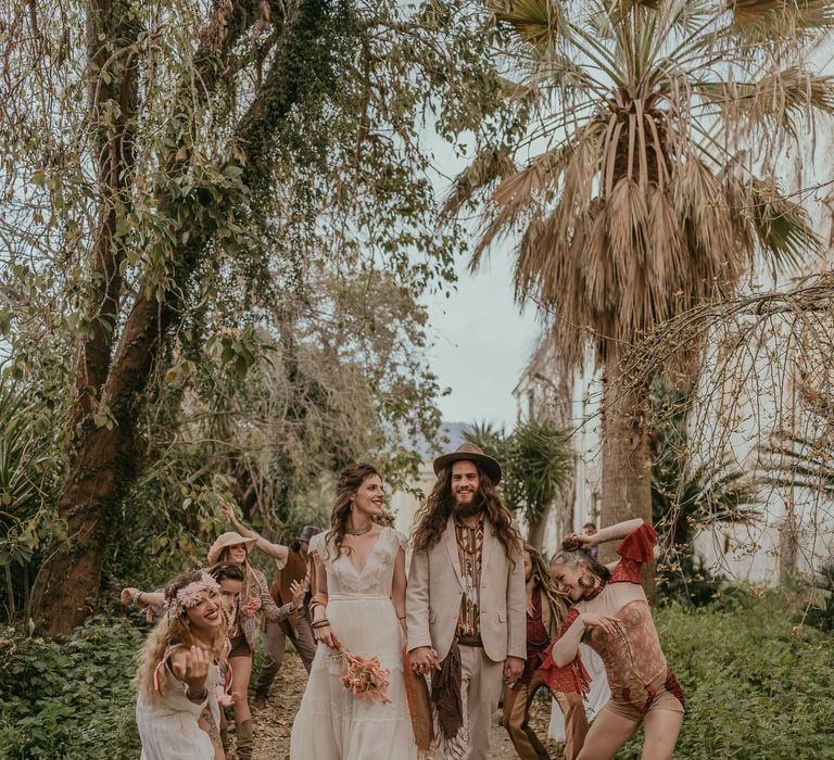 Bride & groom walk with wedding party and circus performers who dance around them as they walk along a country path in Sicily 