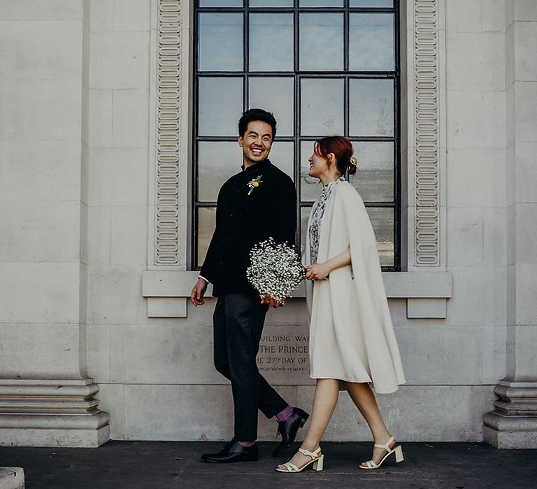 Groom in a blue velvet jacket and bride in a beige cape coat holding a gypsophila wedding bouquet walking outside Old Marylebone Town Hall 