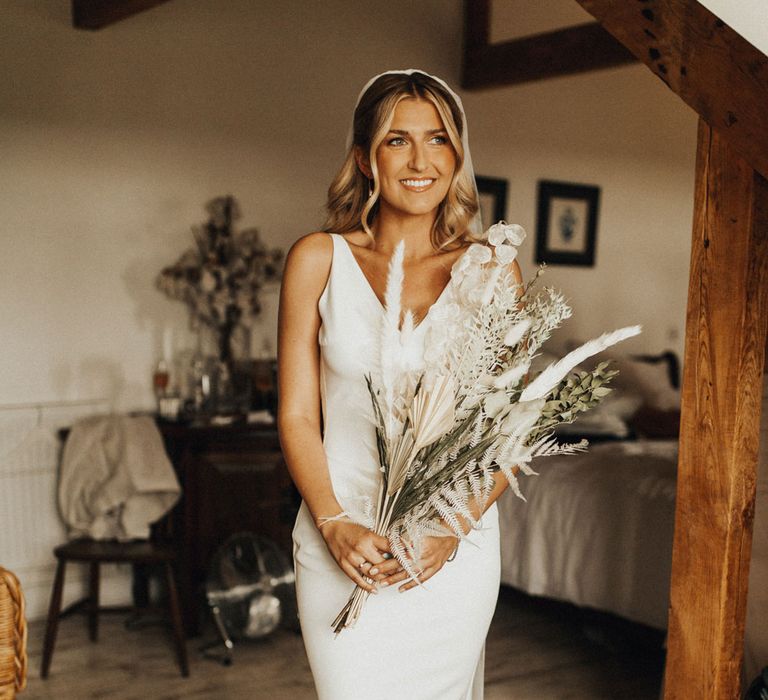 Smiling bride in V neck Made With Love wedding dress and veil holds white and green dried bridal bouquet in bedroom before wedding at Anran Devon