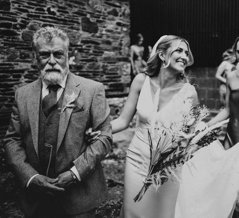 Smiling bride in V neck Made With Love wedding dress and veil holds dried bridal bouquet and arm of man in three piece suit as she walks to get married at Anran Devon