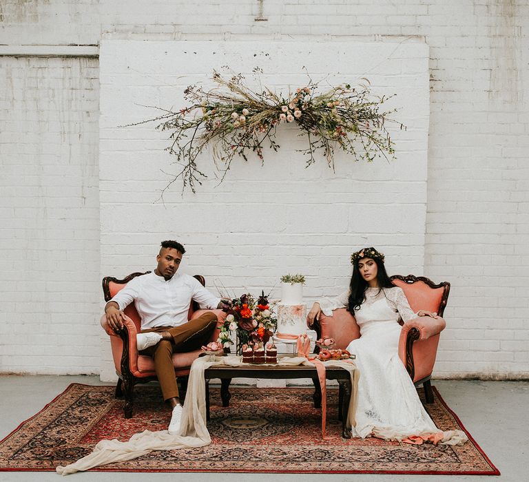 Chill out area with wool rugs, terracotta chairs and a hanging floral installation with boho bride in a lace dress and flower crown and groom in a white grandad shirt 