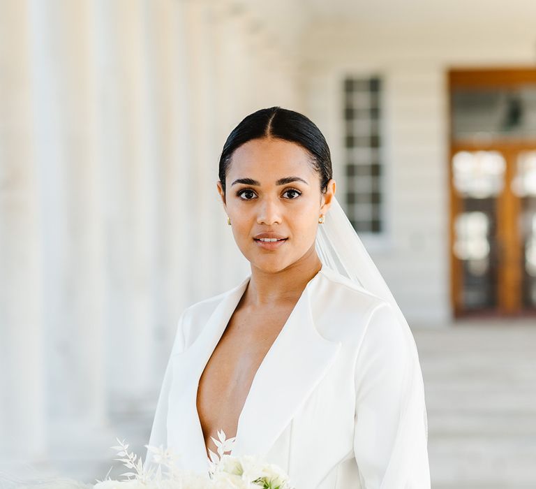 A bride wears a low cut white suit jacket and holds a bouquet filled with white flowers and anemones. She has her hair back with a long veil.