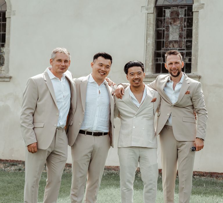 Groom in shawl collared shirt and double breasted linen suit stands with groomsmen in linen suits and white shirts outside church before Wasing Park wedding