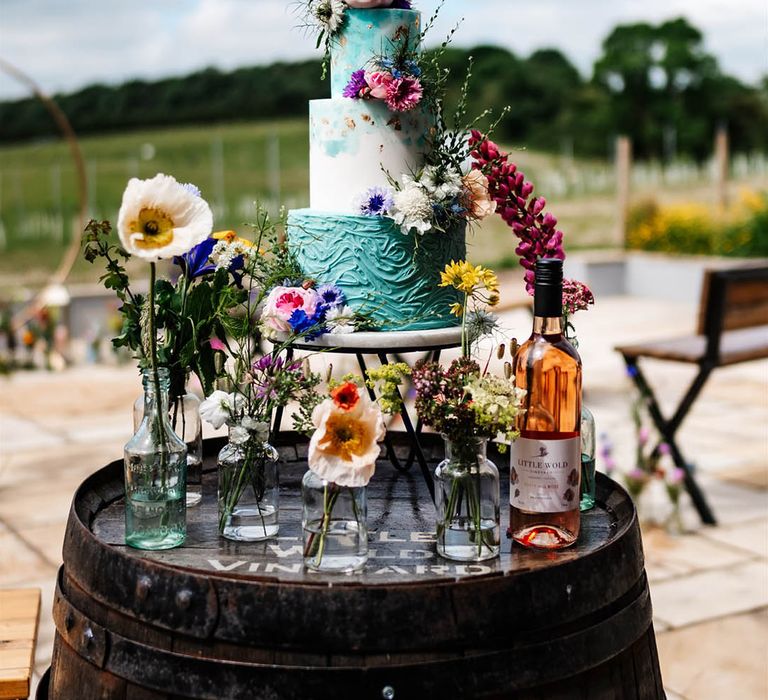 turquoise and white buttercream wedding cake resting on a barrel surrounded by colourful wildflower stems in bottles 