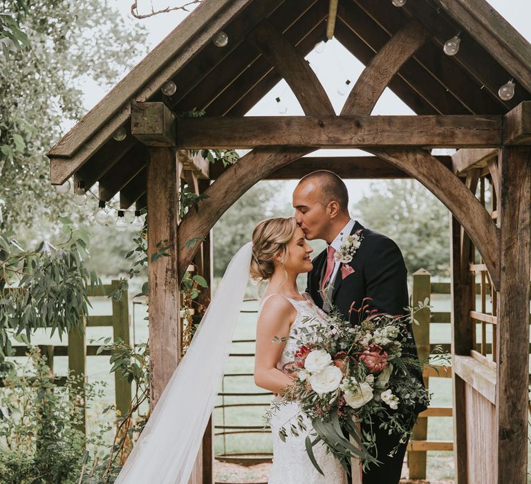 Bride in white lace Justin Alexander wedding dress with train and veil holding white, pink and green bridal bouquet stands by wooden arch as groom in dark three piece suit and pink tie kisses her on the forehead at Tythe Barn wedding with barn wedding flowers