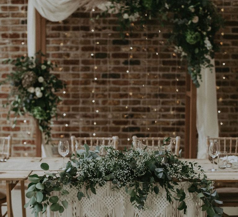 Wooden top table with macrame wedding decor, foliage drapery and bamboo chairs in front of wedding arch with cloth drapery and flower clouds for Isle of Wight wedding with macrame wedding decor