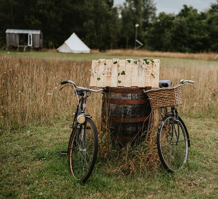 Rustic wooden wedding welcome sign covered in ivy on wooden barrel in-between two bikes for late summer wedding in Norfolk