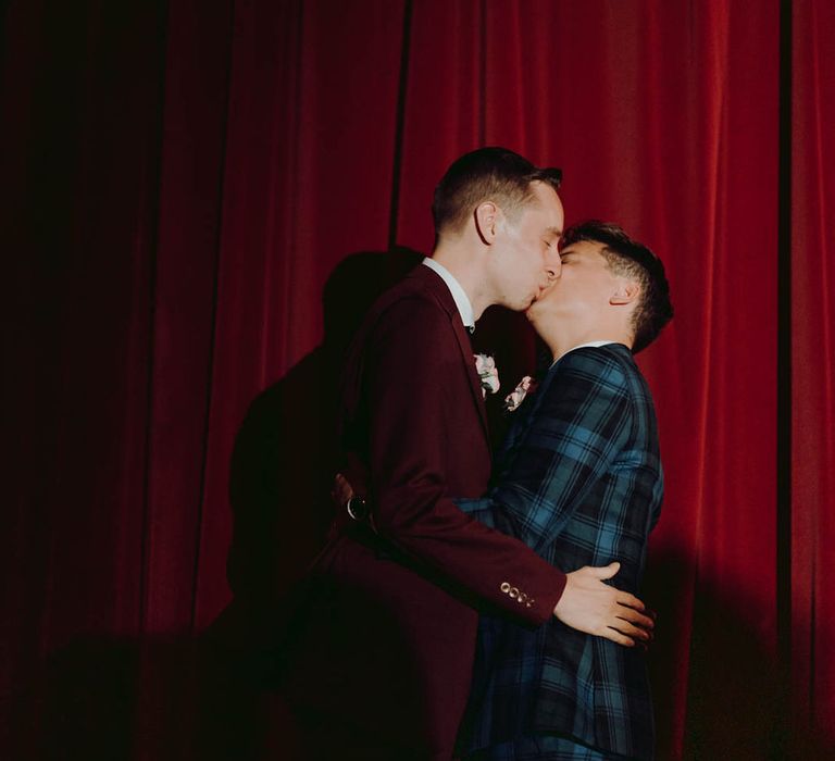 Grooms kiss on the day of their wedding in front of red curtain | Kim Williams Weddings