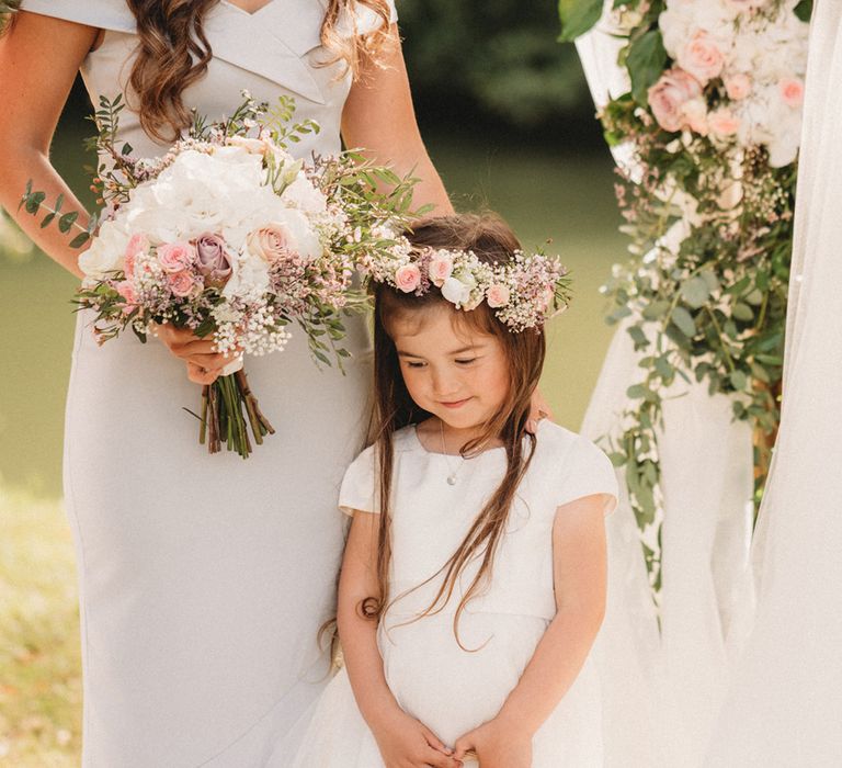 Flower girl in a white dress with white and pink flower crown standing next to the bridesmaid in a light grey dress