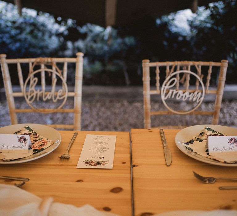 Bride and groom table settings at wooden table with floral stationery, napkins and table place names and bamboo chairs for tipi wedding at Inkersall Grange Farm