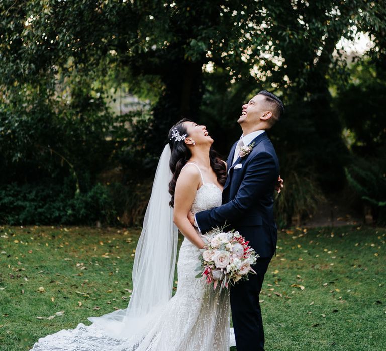 Bride in an embellished wedding dress with thin straps and long train wearing a cathedral length veil and holding a pink protea wedding bouquet laughing with her groom in a navy suit 