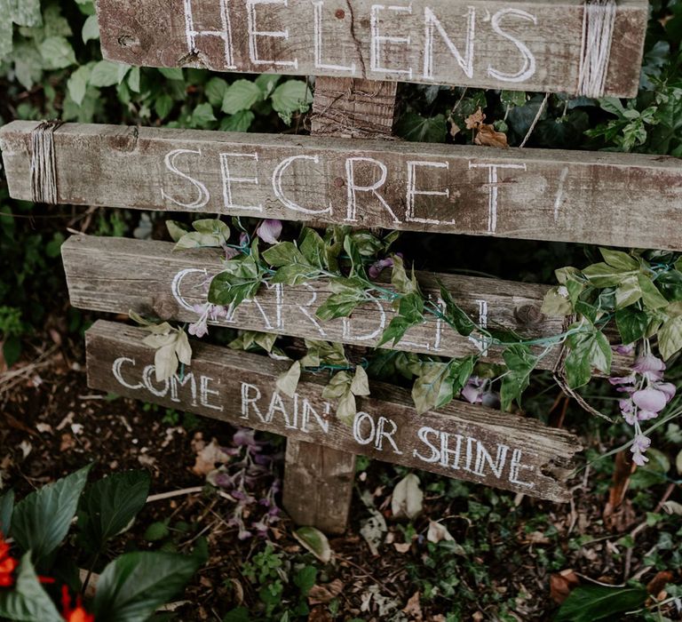 Wooden garden sign with four posts reading 'Helen's Secret Garden Come Rain or Shine' next to bush at DIY garden wedding