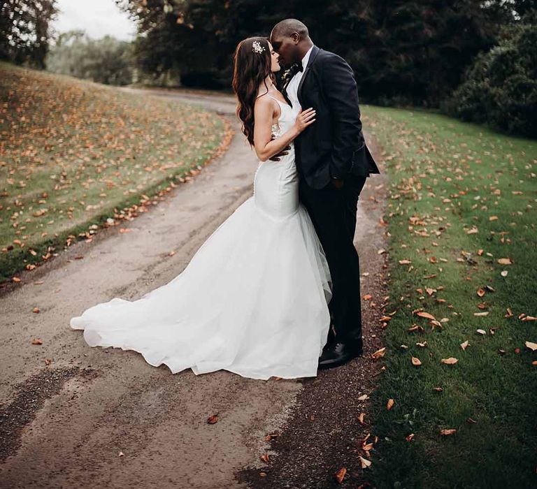 Bride wearing fitted long trail dress kissing Groom in dark fitted suit outdoors with autumnal leafs on the ground