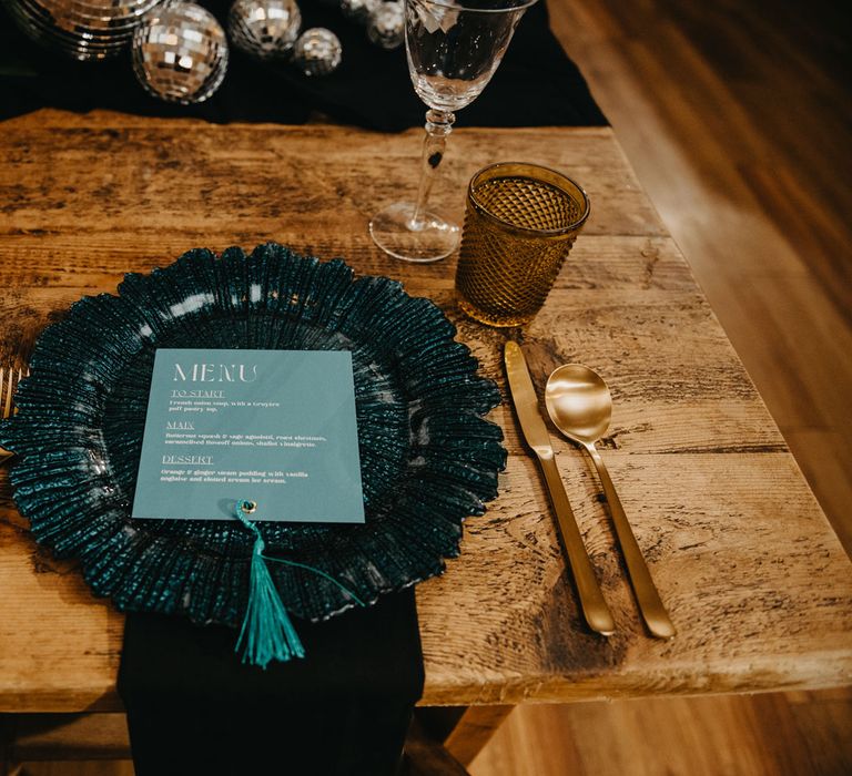 Blue glass plate with light blue wedding menu on wooden table with gold cutlery and orange glasses for rustic wedding breakfast