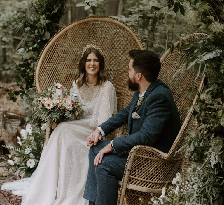 Bride & groom look lovingly at one another whilst they sit in wicker sweetheart chairs