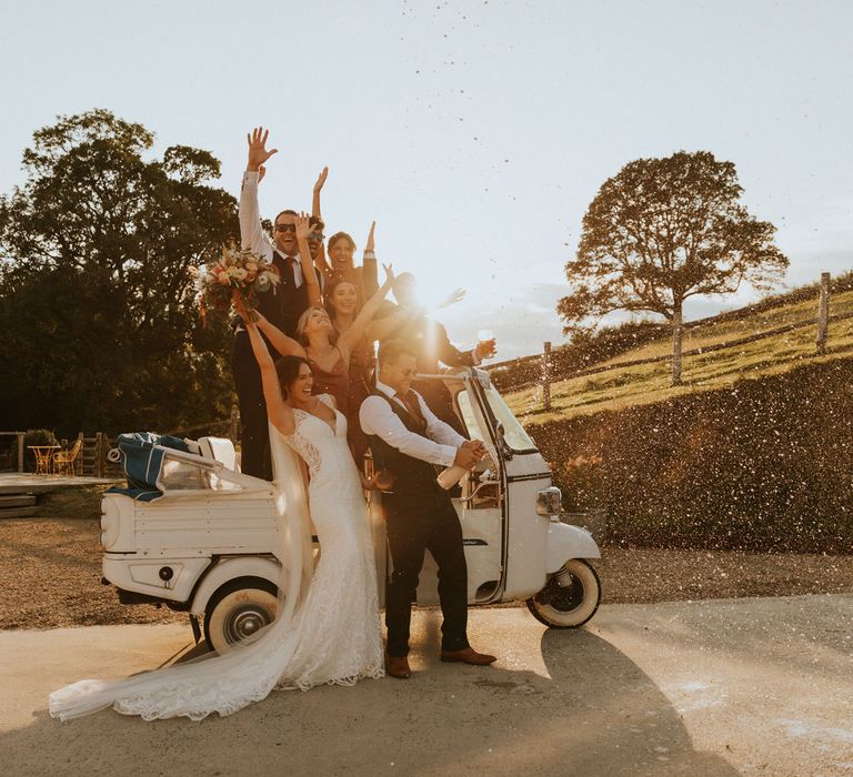 Bride in lace wedding dress with Tain stands by cart with groom and wedding guests as groom sprays champagne during golden hour