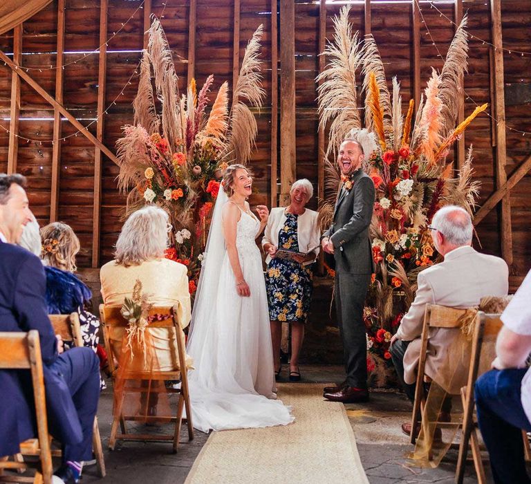 Bride in lace thin strap wedding dress and veil stands with groom in green three piece suit in front of rustic orange pampas grass and rose floral installations during wedding ceremony