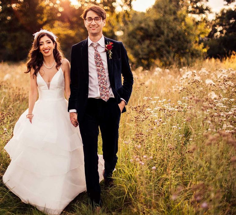 Smiling bride in wedding dress with tulle skirt and plunging neckline walks through field with groom in dark suit, Gucci tie and floral buttonhole during golden hour