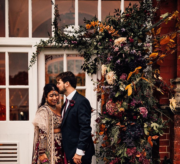 Bride in Indian Saree wedding dress stands with groom under their flower arch full of autumnal colours 