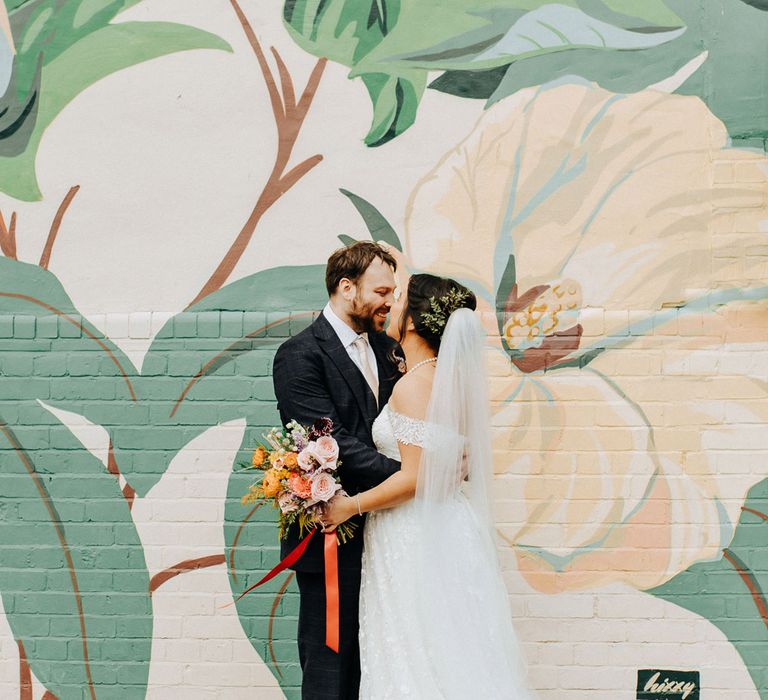 Bride in lace wedding dress with train and veil holding bouquet tied with red ribbon stands with groom in blue check suit in front of wall covered in street art in Liverpool