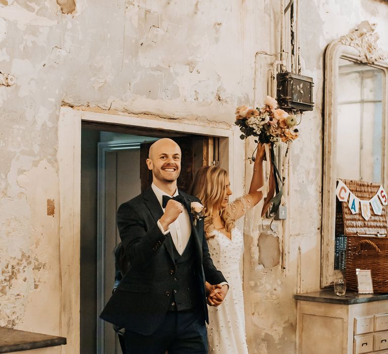 Groom in navy three piece tweed suit with bow tie walks into wedding reception with bride in white pearl wedding dress holding up pastel wedding bouquet