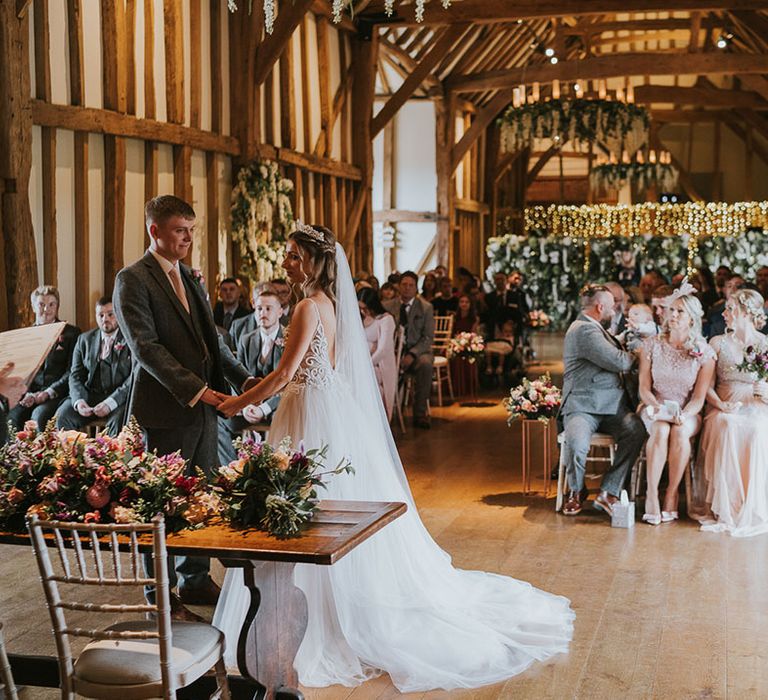 Bride and groom at the altar with bride in tulle puddle train dress at rustic wedding venue complete with hanging wisteria and fairy lights 