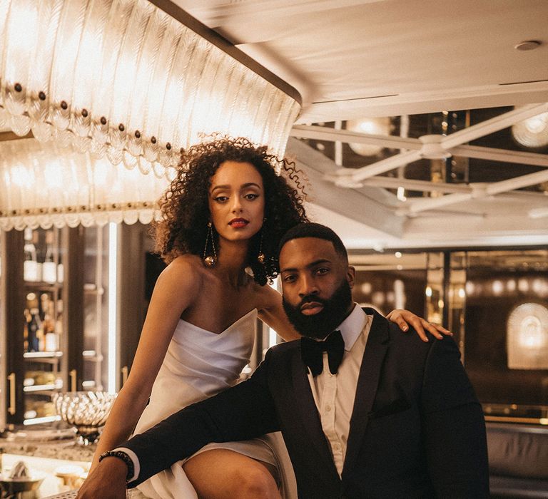 Black groom in a tuxedo standing at the bar with his Black bride in a strapless wedding dress with front slit sitting on the bar with naturally curly hair, red lipstick and winged eyeliner makeup 