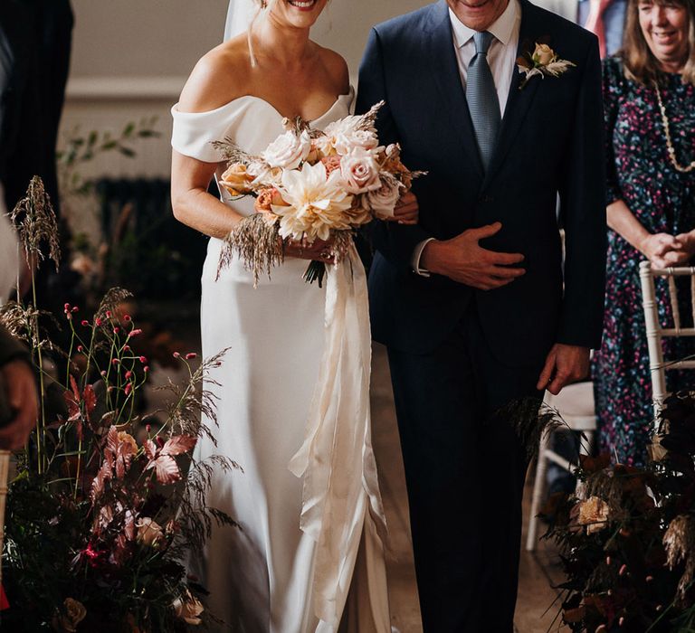 Father of the bride in blue suit walks the bride down the aisle wearing an off the shoulder front slit wedding dress