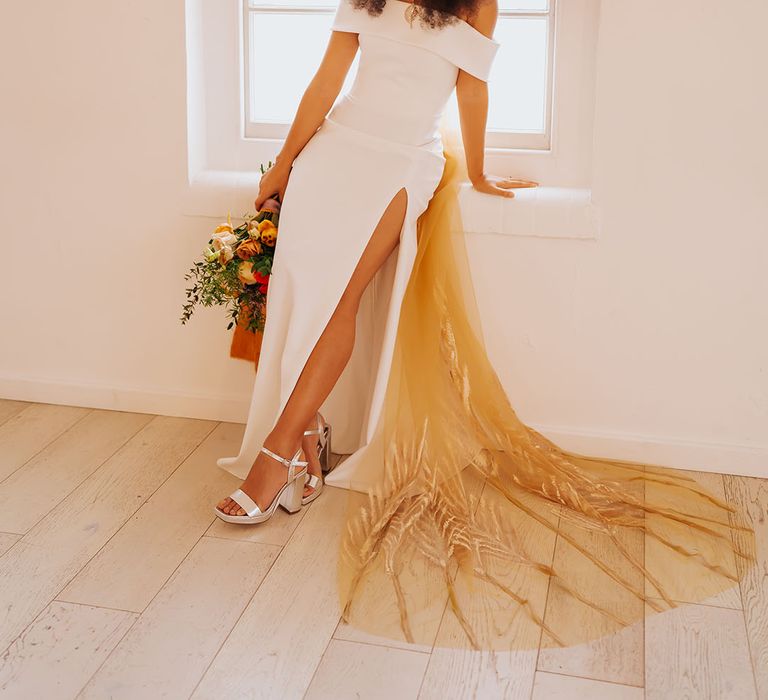 Black bride in an off the shoulder wedding dress with side slit wearing silver platform shoes and a patterned golden wedding veil