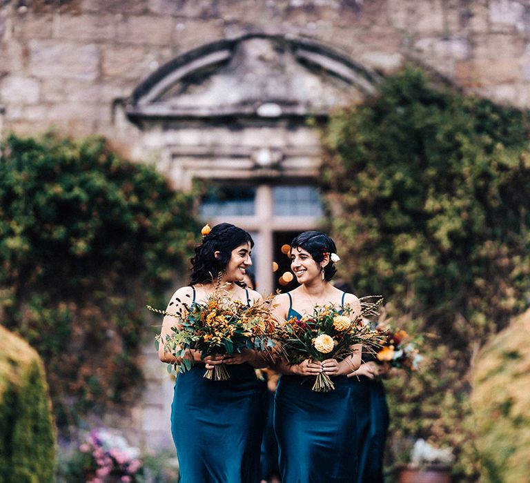 Bridesmaids in blue dresses smile at each other as they walk down the aisle holding autumnal bouquets 