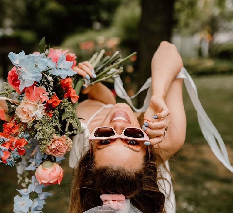 Bride in retro pink sunglasses holding a pink and blue wedding veil with peonies and anthuriums 