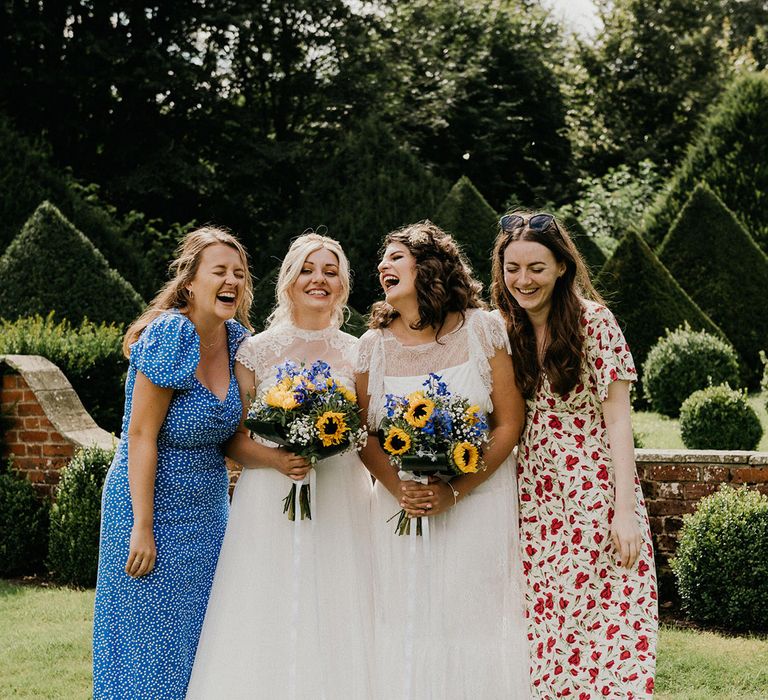 Brides laugh together with two wedding guests in flower and polka dot dresses as they hold their sunflower and gypsophila bouquets