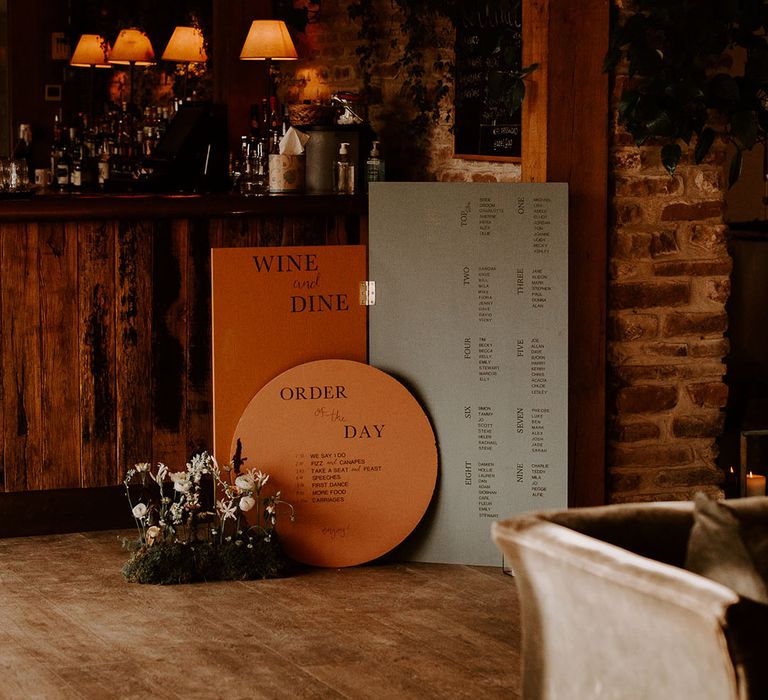 Circular orange order of the day sign next to green and orange seating chart with 'wine and dine' sign and white flowers