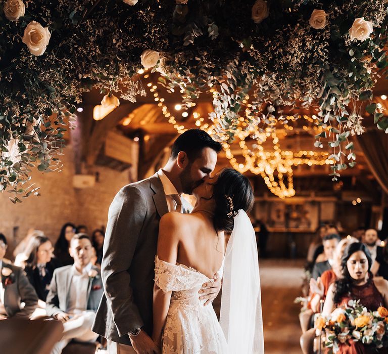 Bride in lace off the shoulder wedding dress and groom in grey suit kiss under flower display for their barn wedding 