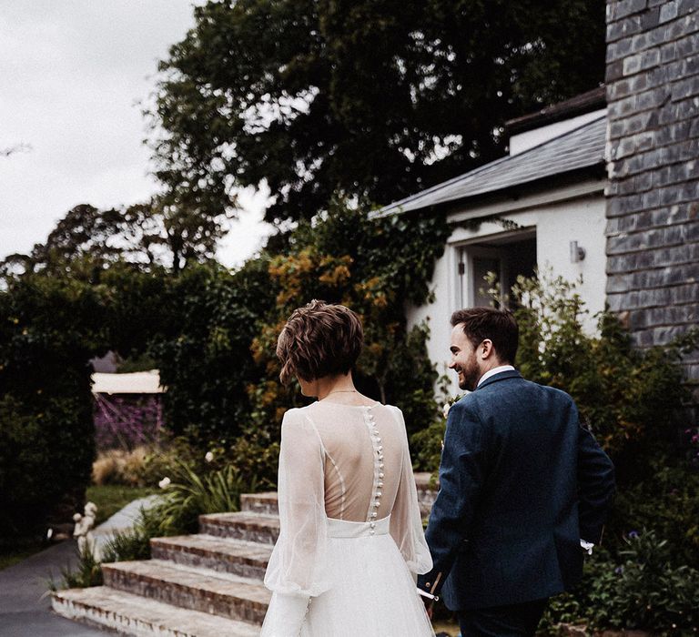 Groom in blue suit walks holding hands with bride in sheer long sleeve wedding dress 