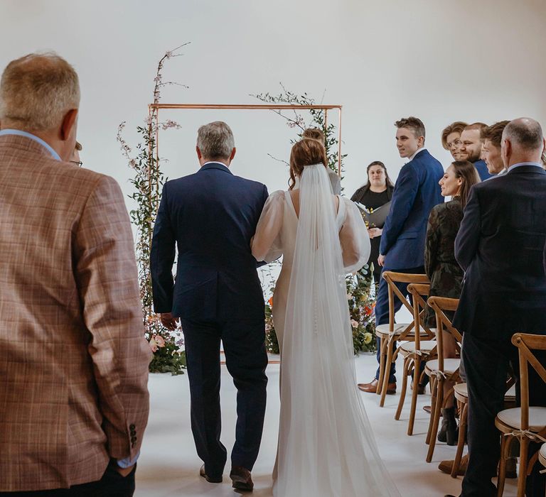 Bride in wedding dress with button detail and long veil is walked down there aisle by father in blue suit to altar with copper frame and foliage