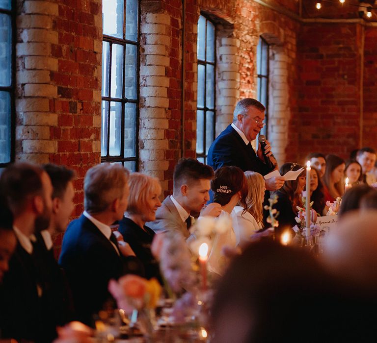 Father of the bride in navy blue suit reads out his speech at wedding decorated with candle light and fairy lights 