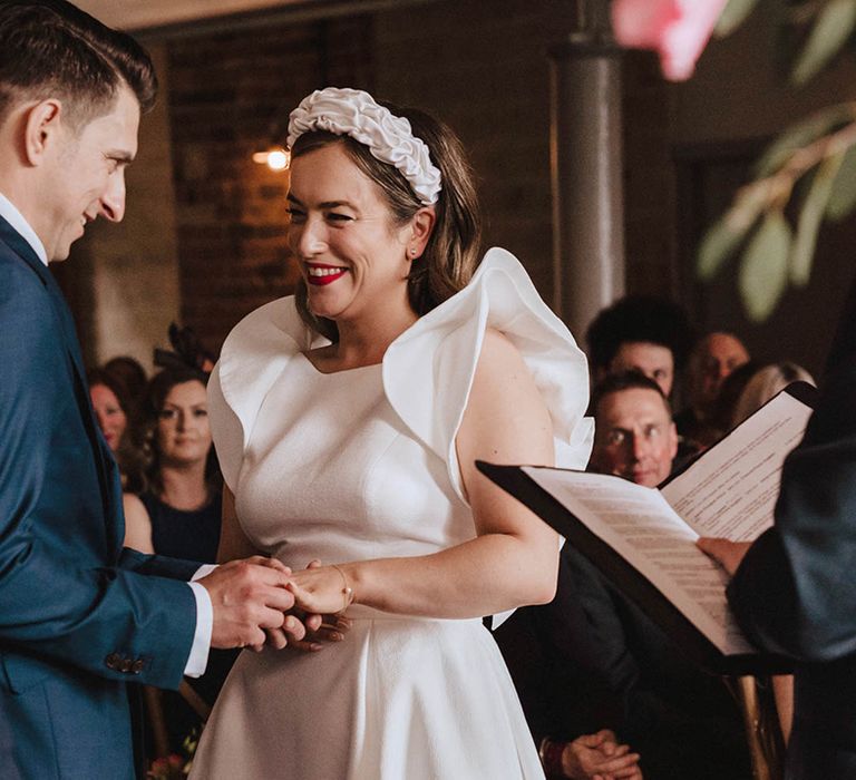 Bride in ruffle sleeve Jesus Peiro wedding dress and large headband with red lipstick smiles as the groom puts a ring on her finger