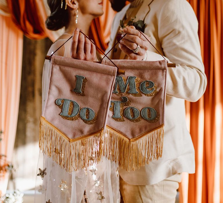 Bride in a celestial wedding dress and groom in a beige suit holding pink banners saying 'I do' and 'me too'