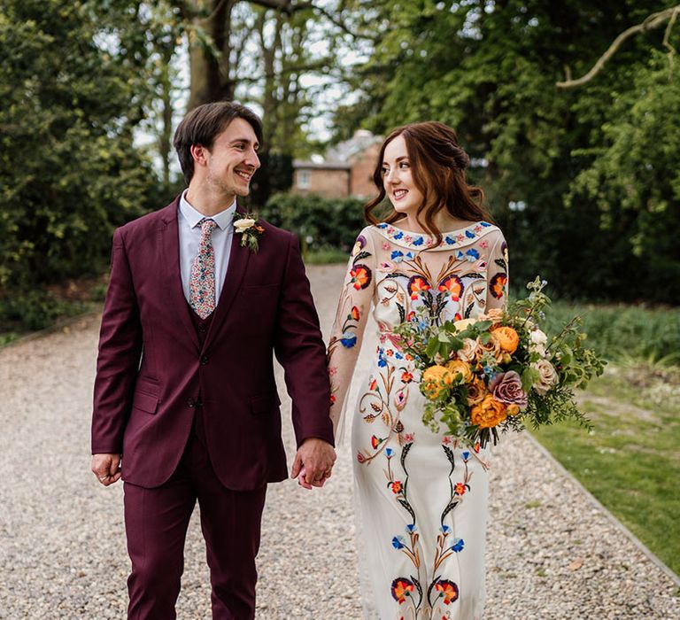 Groom in three piece burgundy suit with bride in colourful flower wedding dress with colourful bouquet 