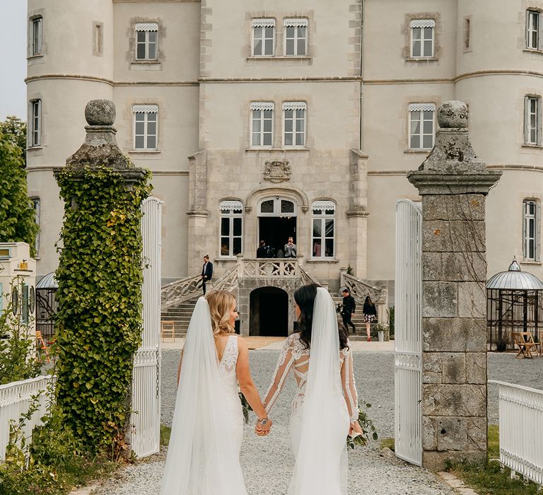 Brides walk through white iron gate together holding hands in front of the chateau