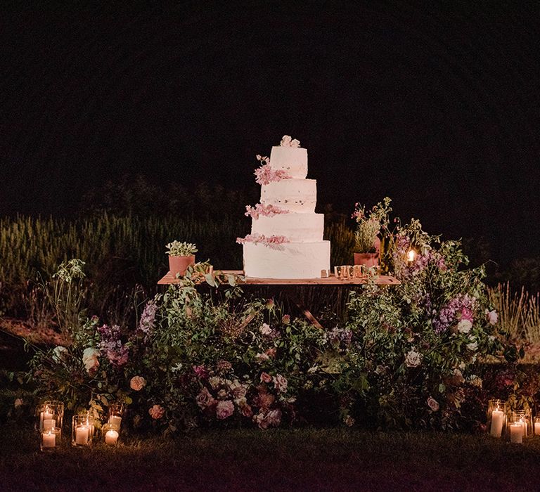 Four tier rustic wedding cake complete with pale pink floral decor sits on wooden table outdoors surrounded by florals and green foliage