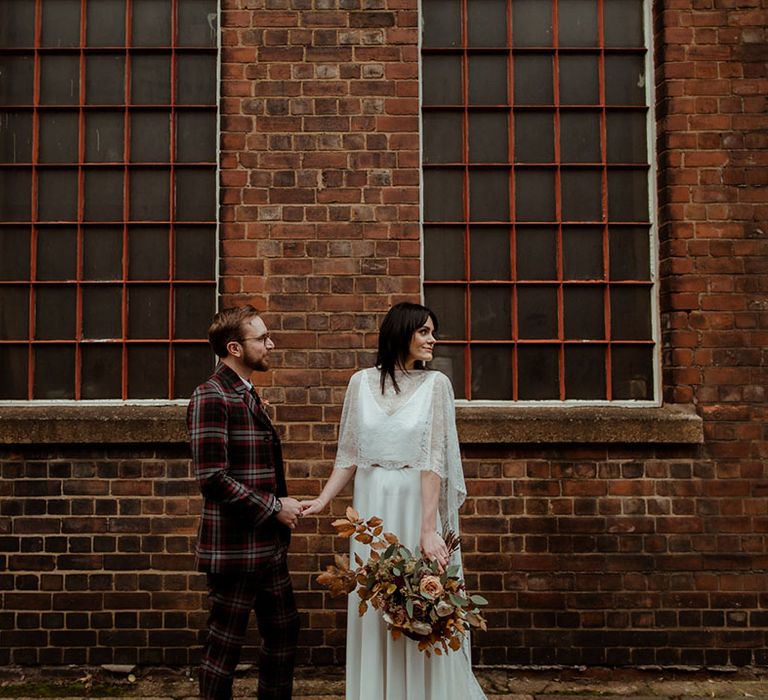 Bride & groom stand in front of industrial setting at The Chimney House as bride wears lace cape and groom wears bespoke tartan suit