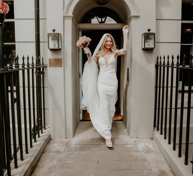 Bride walks out in her Made With Love wedding dress and bridal gloves with pink bouquet out of the townhouse ready for her wedding 