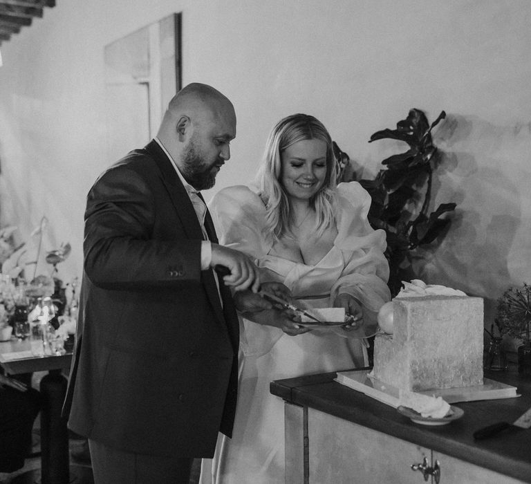 black and white portrait of the bride in a wedding dress with long puffy sleeves cutting the square wedding cake 