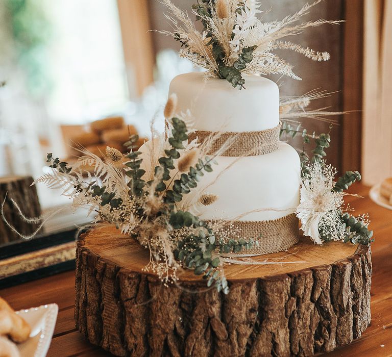 Simple two tier iced wedding cake with dried grasses for a boho effect on a tree slice cake stand 