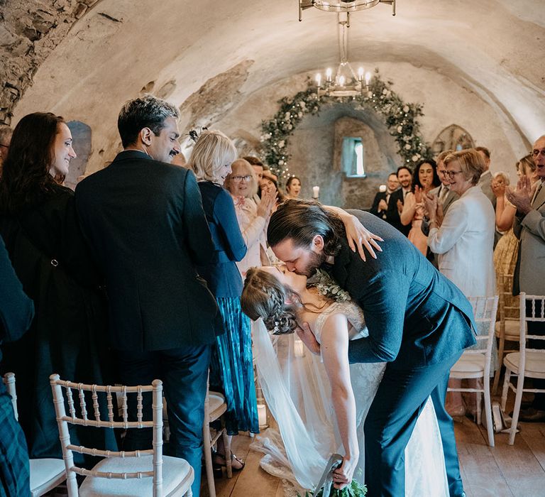 Groom dips his bride for a kiss in rustic styled room in Neidpath Castle complete with chandelier and floral archway 