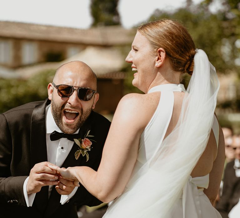 Groom wearing sunglasses and black tie with floral buttonhole puts wedding ring on his bride during outdoor ceremony in France