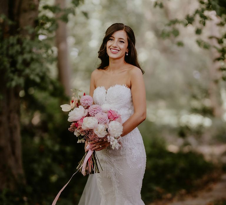 Bride wears lace Pronovias wedding dress with sweetheart neckline and her brown hair in loose waves whilst holding white and pink floral bouquet