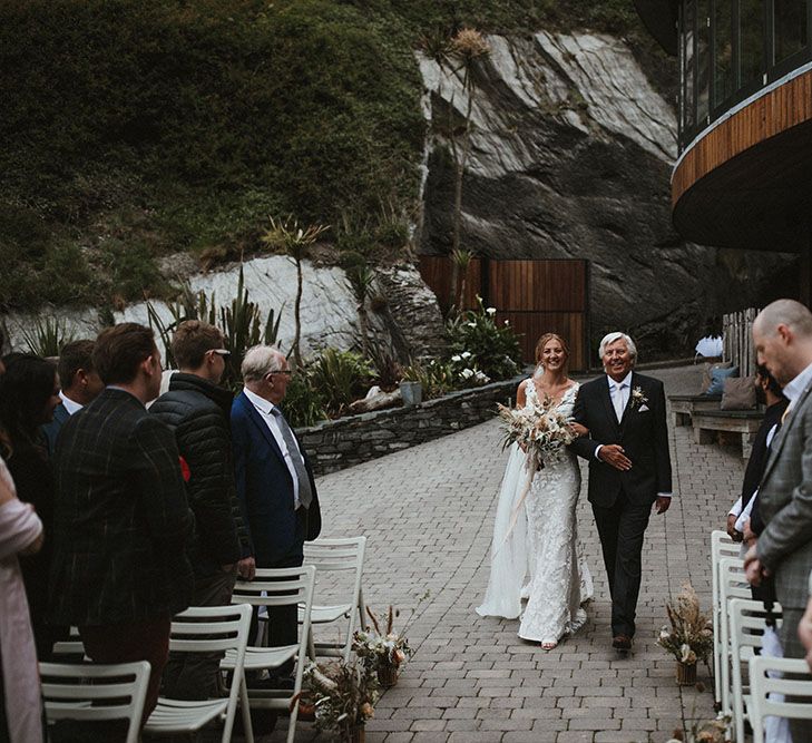 Bride wears lace Enzoani wedding dress as she walks down the aisle with her father who wears black suit and floral buttonhole 